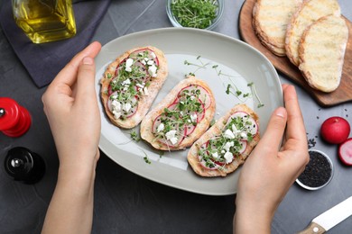 Photo of Woman holding delicious sandwiches with radish, cheese and microgreens at table, top view