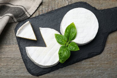 Photo of Tasty cut and whole brie cheeses with basil on wooden table, flat lay
