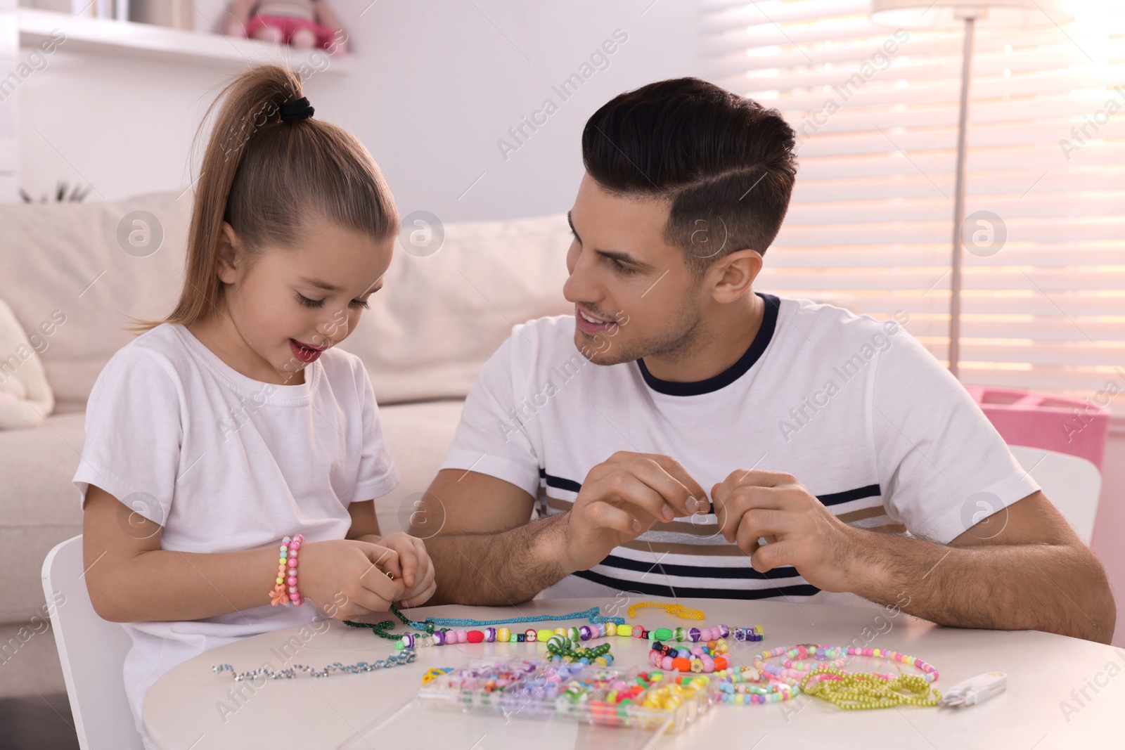 Photo of Happy father with his cute daughter making beaded jewelry at table in room