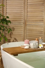 Photo of Wooden tray with spa products, stones and burning candle on bath tub in bathroom. Space for text