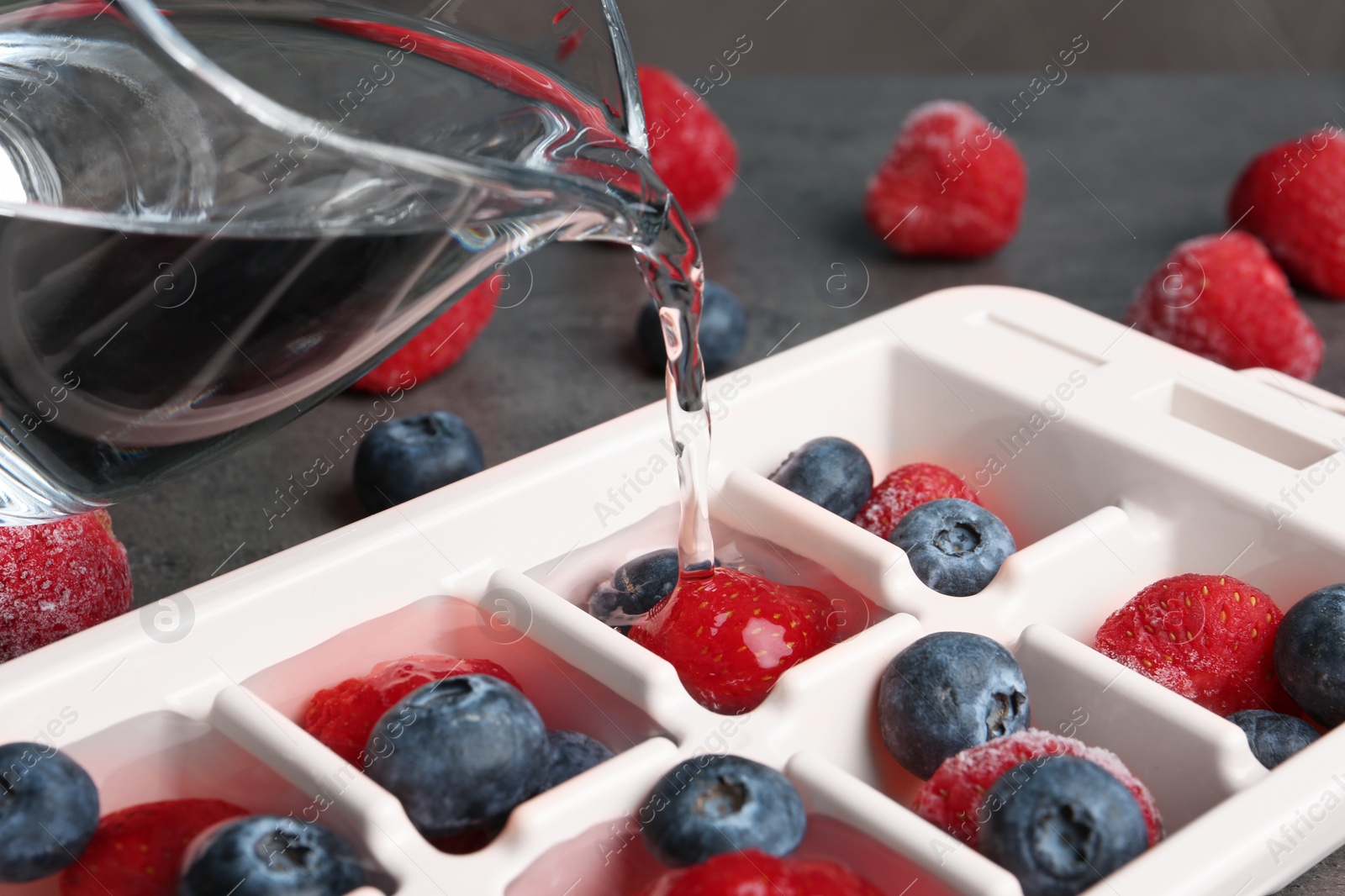 Photo of Pouring water into ice cube tray with berries on table, closeup