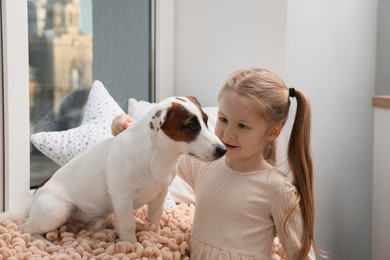 Photo of Cute little girl with her dog near window at home. Childhood pet