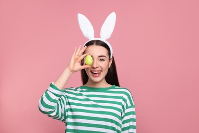 Happy woman in bunny ears headband holding painted Easter egg on pink background