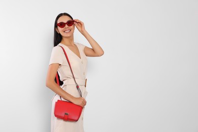 Beautiful young woman with red stylish bag in sunglasses on light grey background, space for text