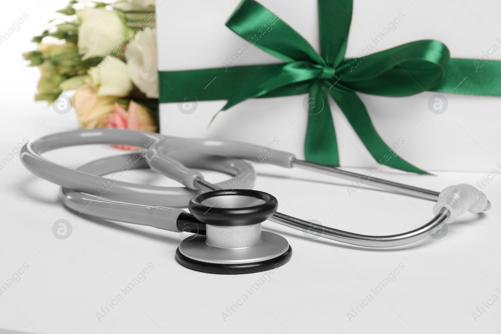 Photo of Stethoscope, gift box and eustoma flowers on white background, closeup. Happy Doctor's Day