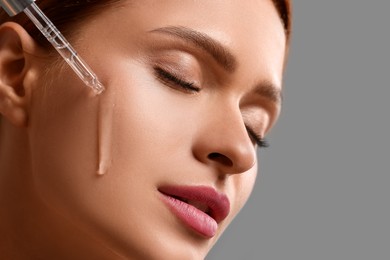 Photo of Beautiful young woman applying cosmetic serum onto her face on grey background, closeup