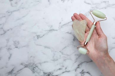 Woman holding gua sha stone and face roller at white marble table, top view. Space for text