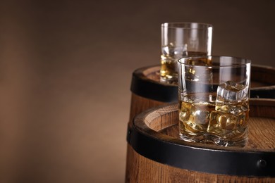 Photo of Whiskey with ice cubes in glasses on wooden barrels against brown background, closeup. Space for text