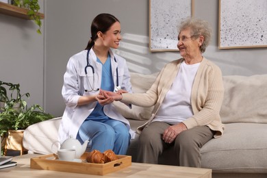 Photo of Young caregiver examining senior woman on sofa in room. Home health care service