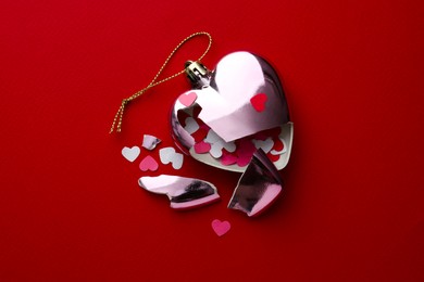 Crashed bauble in shape of heart with confetti on red background, top view. Broken heart