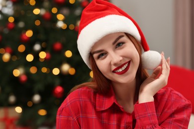 Beautiful young woman in Santa hat near Christmas tree indoors, space for text