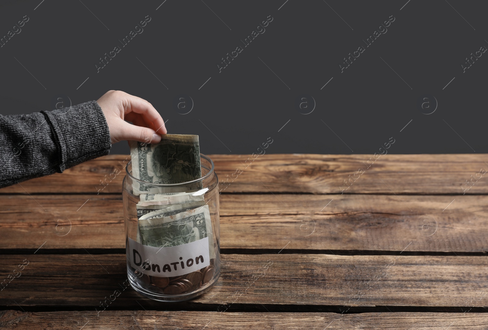 Photo of Woman putting money into donation jar on wooden table against grey background, closeup. Space for text