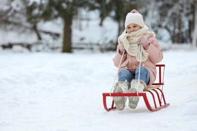 Photo of Cute little girl enjoying sledge ride through snow in winter park, space for text