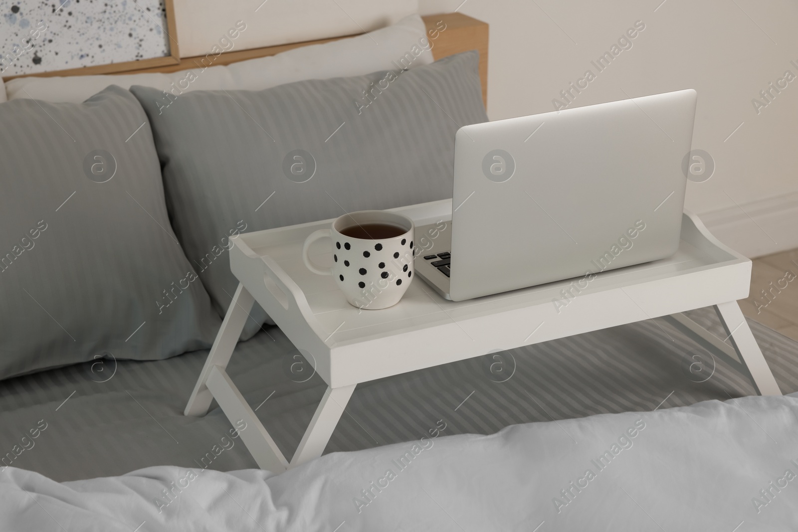 Photo of White tray table with laptop and cup of drink on bed indoors