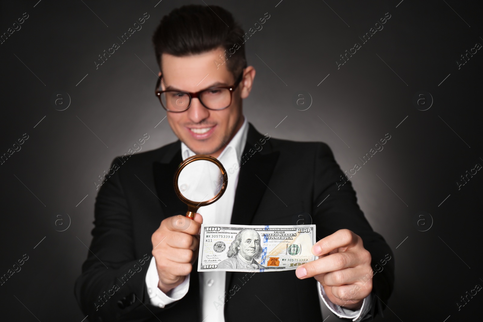 Photo of Expert authenticating 100 dollar banknote with magnifying glass against dark background, focus on hand. Fake money concept