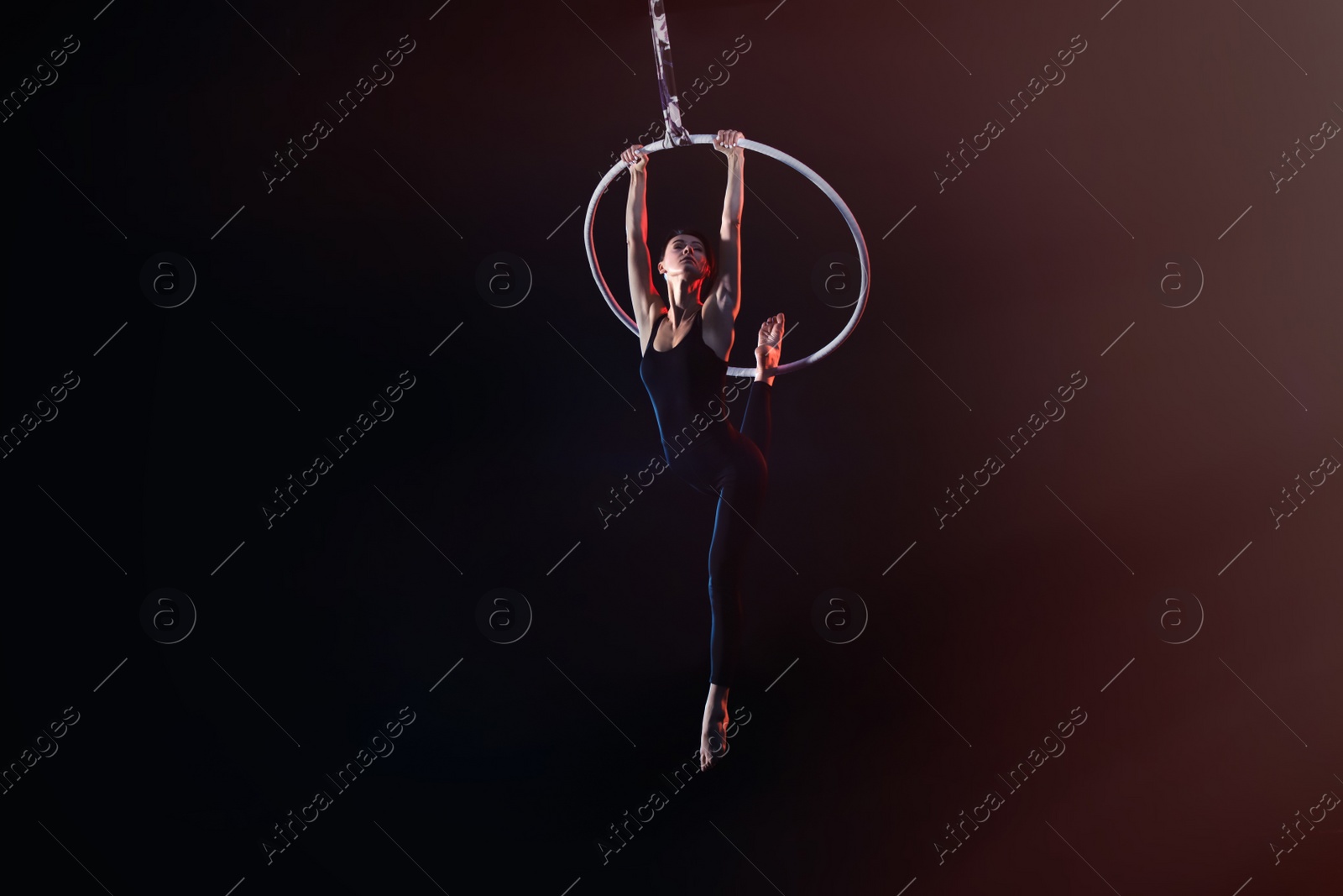 Photo of Young woman performing acrobatic element on aerial ring against dark background