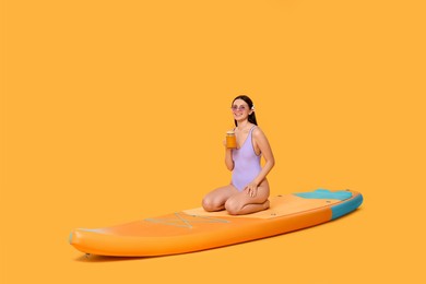 Happy woman with refreshing drink resting on SUP board against orange background