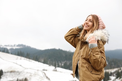 Young woman in warm clothes near snowy hill, space for text. Winter vacation