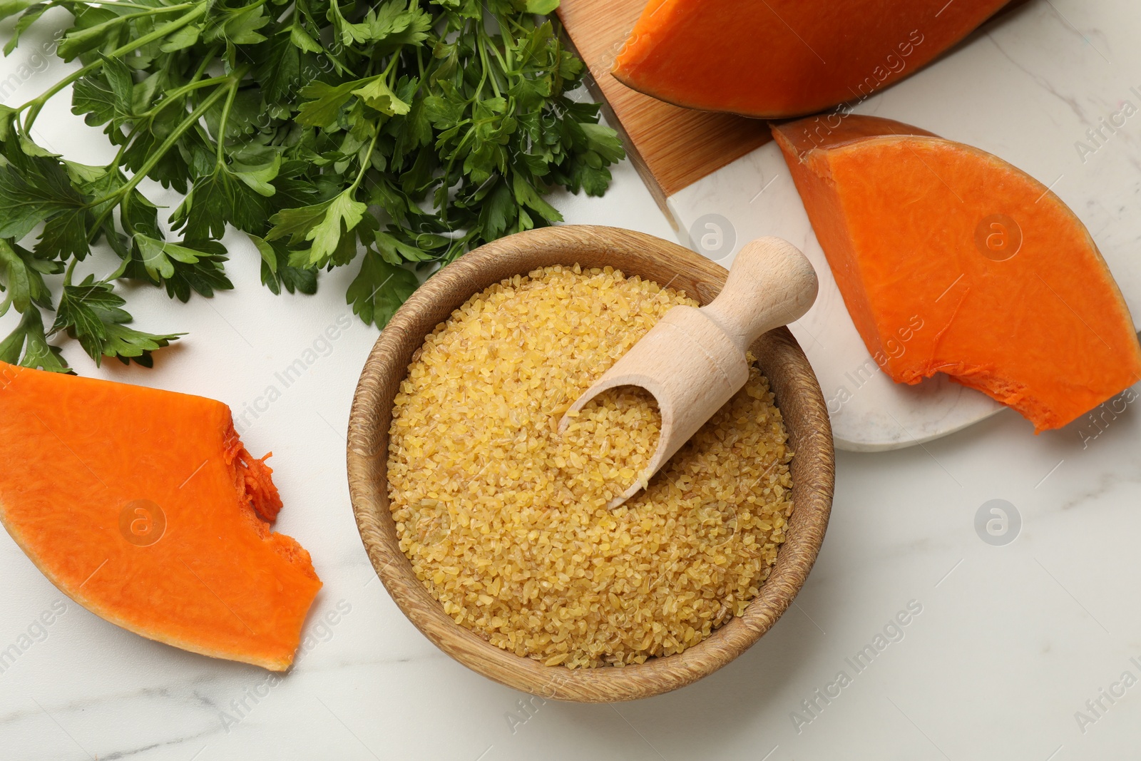 Photo of Raw bulgur in bowl, scoop, pieces of pumpkin and parsley on white table, flat lay