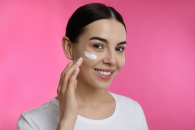 Photo of Young woman applying facial cream on pink background