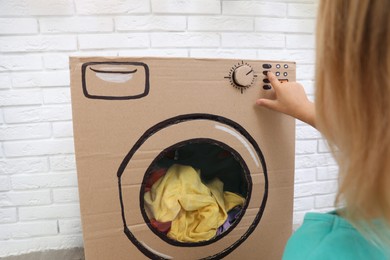 Photo of Little girl playing with toy cardboard washing machine indoors, closeup