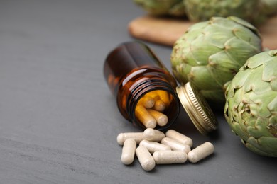 Photo of Bottle with pills and fresh artichokes on grey wooden table, closeup. Space for text
