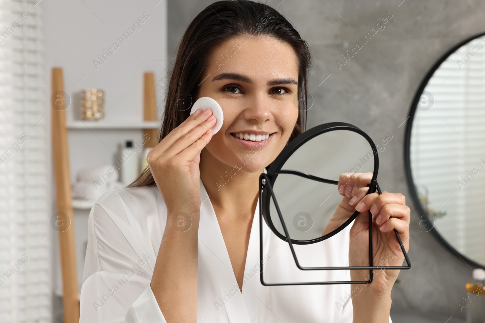 Photo of Young woman cleaning her face with cotton pad near mirror in bathroom