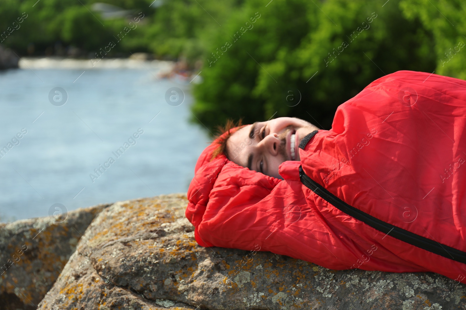 Photo of Young man resting in red sleeping bag near lake. Space for text