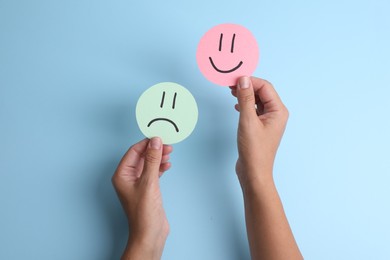 Photo of Choice concept. Woman holding papers with sad and happy emoticons on light blue background, top view