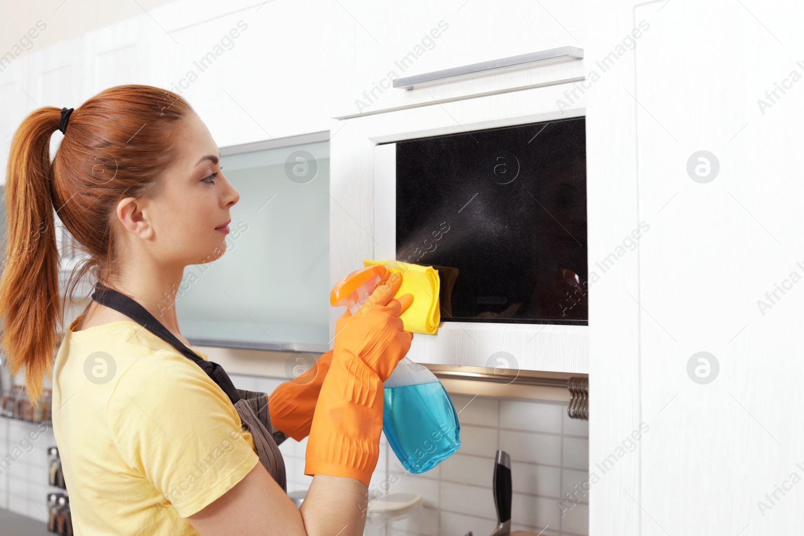 Photo of Woman cleaning microwave oven with rag and detergent in kitchen