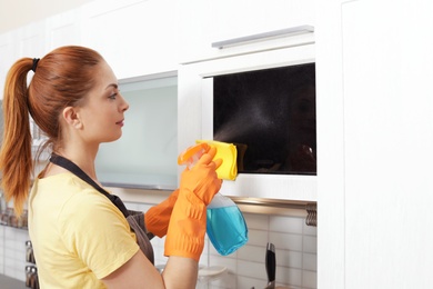 Woman cleaning microwave oven with rag and detergent in kitchen