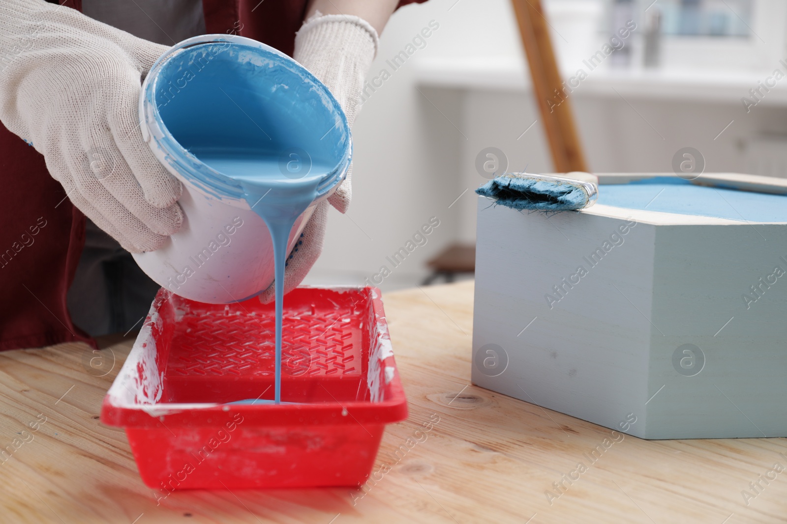 Photo of Woman pouring light blue paint from bucket into tray at wooden table indoors, closeup