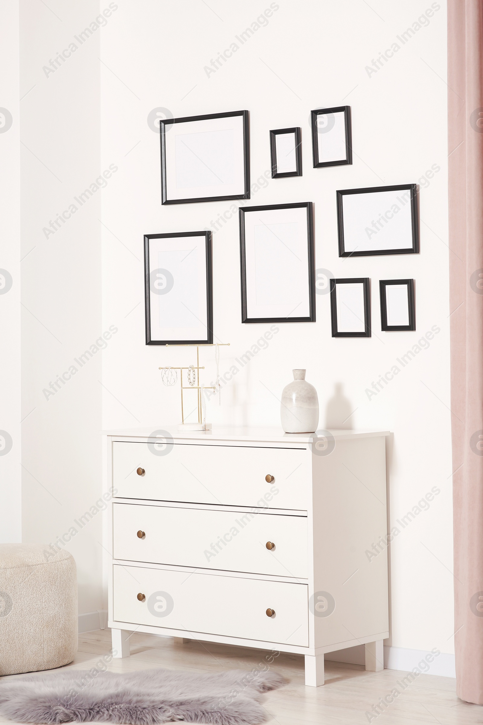 Photo of Empty frames hanging on white wall, chest of drawers and ottoman indoors