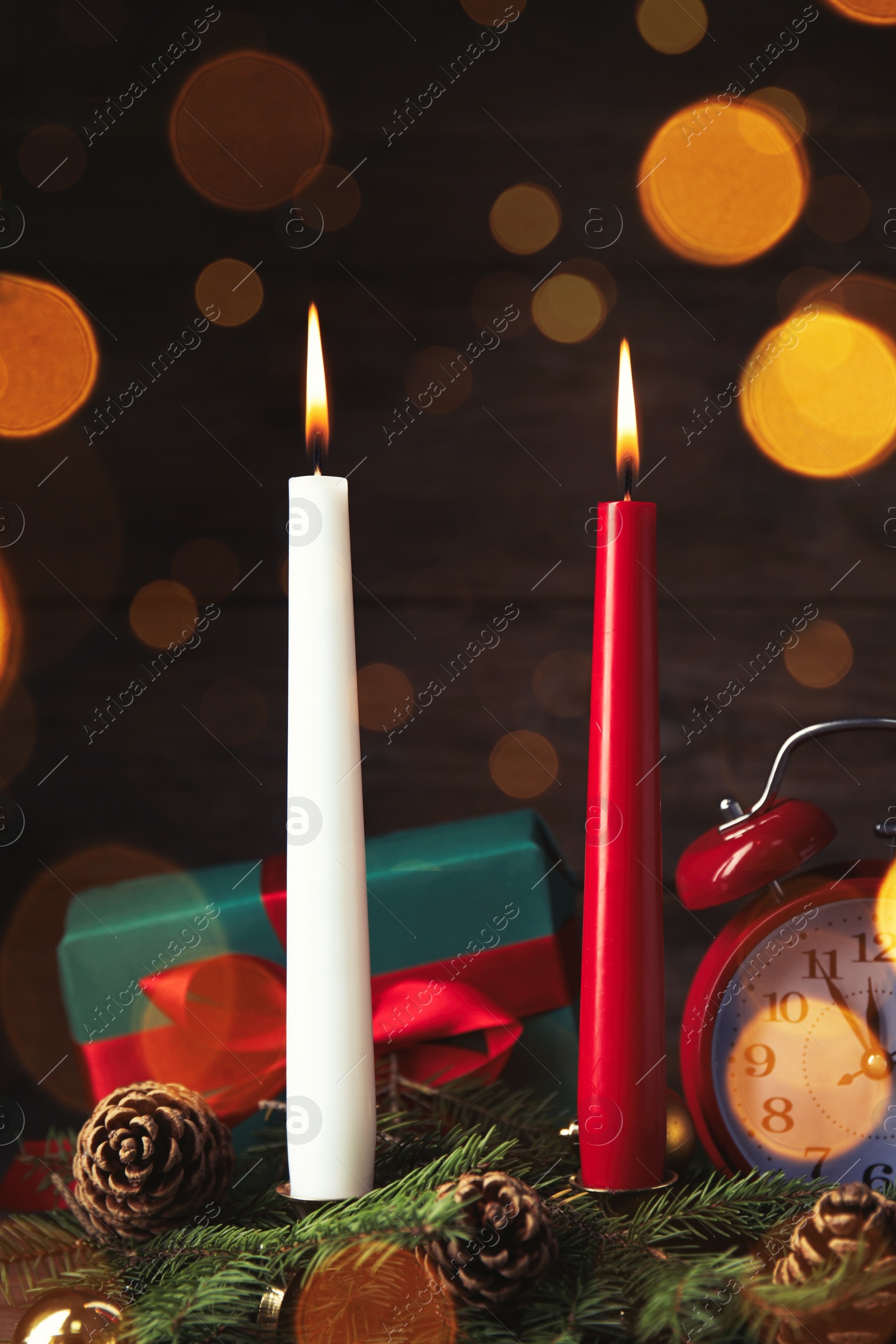 Photo of Burning candles, fir branches, gift box and alarm clock on wooden table, bokeh effect