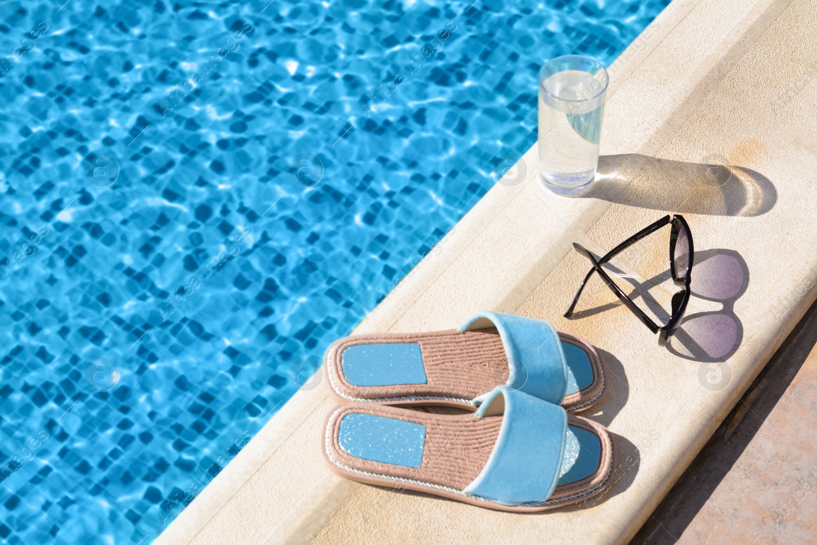 Photo of Stylish sunglasses, slippers and glass of water at poolside on sunny day, space for text. Beach accessories