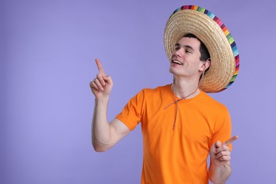 Young man in Mexican sombrero hat pointing at something on violet background. Space for text