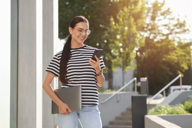 Happy young woman with modern laptop using smartphone outdoors. Space for text