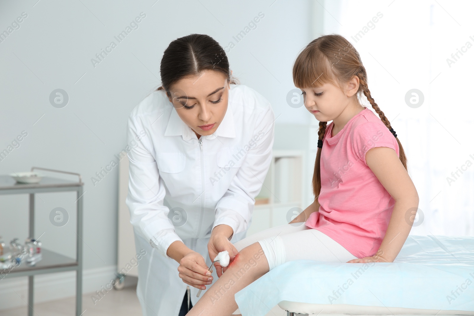 Photo of Female doctor cleaning little girl's leg injury in clinic. First aid