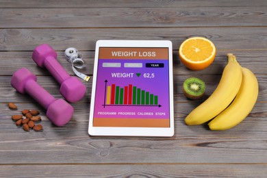 Tablet with weight loss calculator application, dumbbells and products on grey wooden table