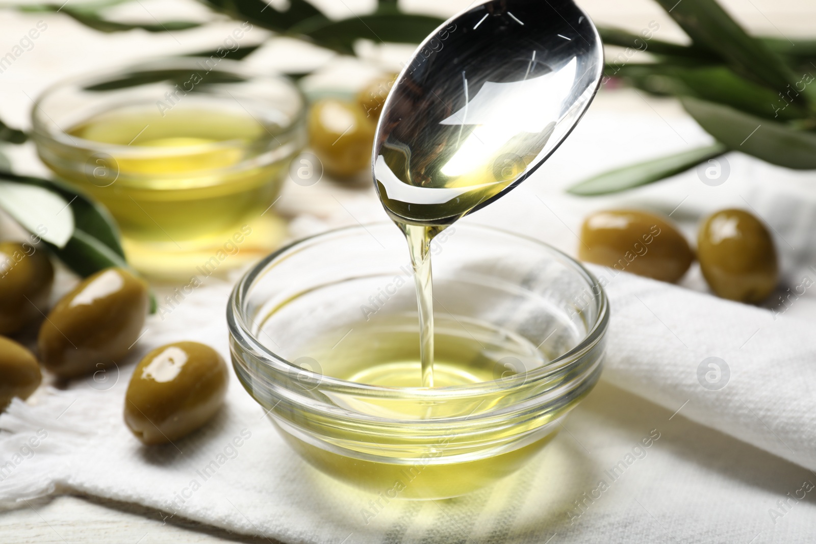 Photo of Spoon with cooking oil over bowl and olives on white wooden table, closeup