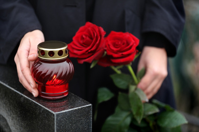 Photo of Woman holding red roses near black granite tombstone with candle outdoors, closeup. Funeral ceremony