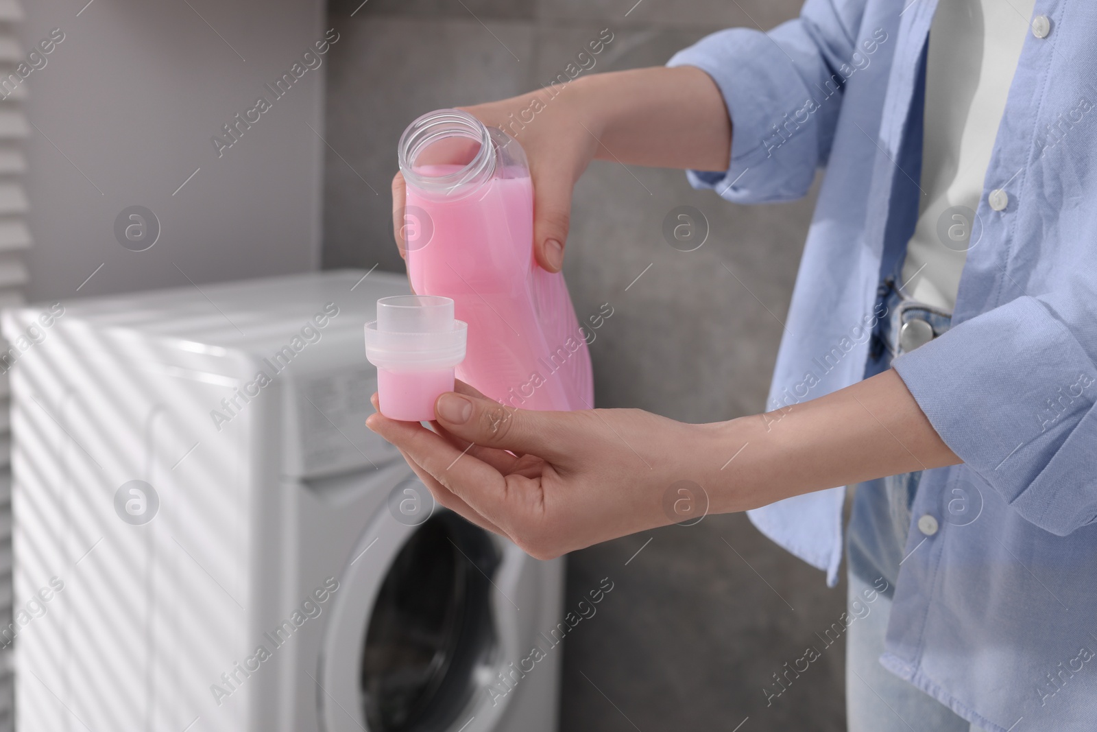 Photo of Woman pouring fabric softener from bottle into cap near washing machine indoors, closeup