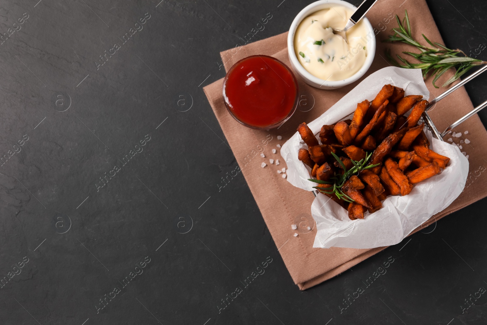 Photo of Frying basket with sweet potato fries and sauces on black table, top view. Space for text