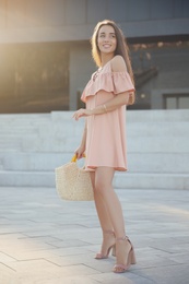 Beautiful young woman in stylish pink dress with handbag on city street