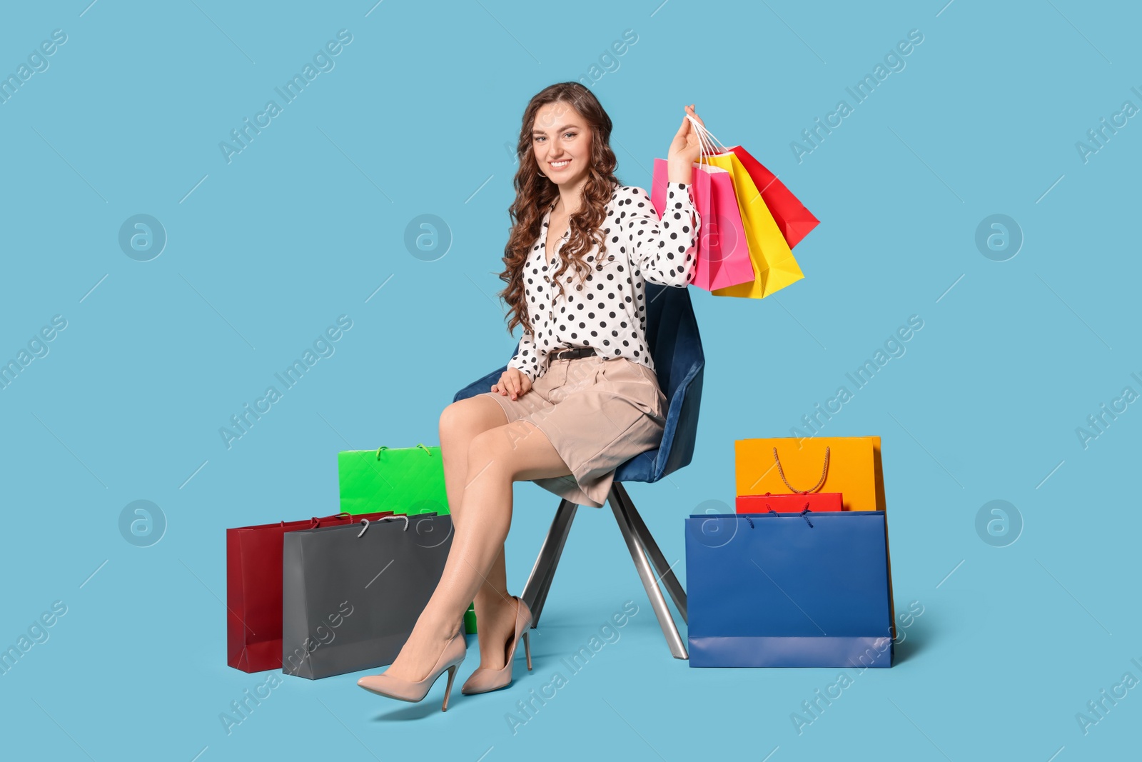 Photo of Happy woman holding colorful shopping bags on armchair against light blue background