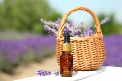 Photo of Bottle of essential oil and wicker bag with lavender flowers on white wooden surface outdoors, closeup