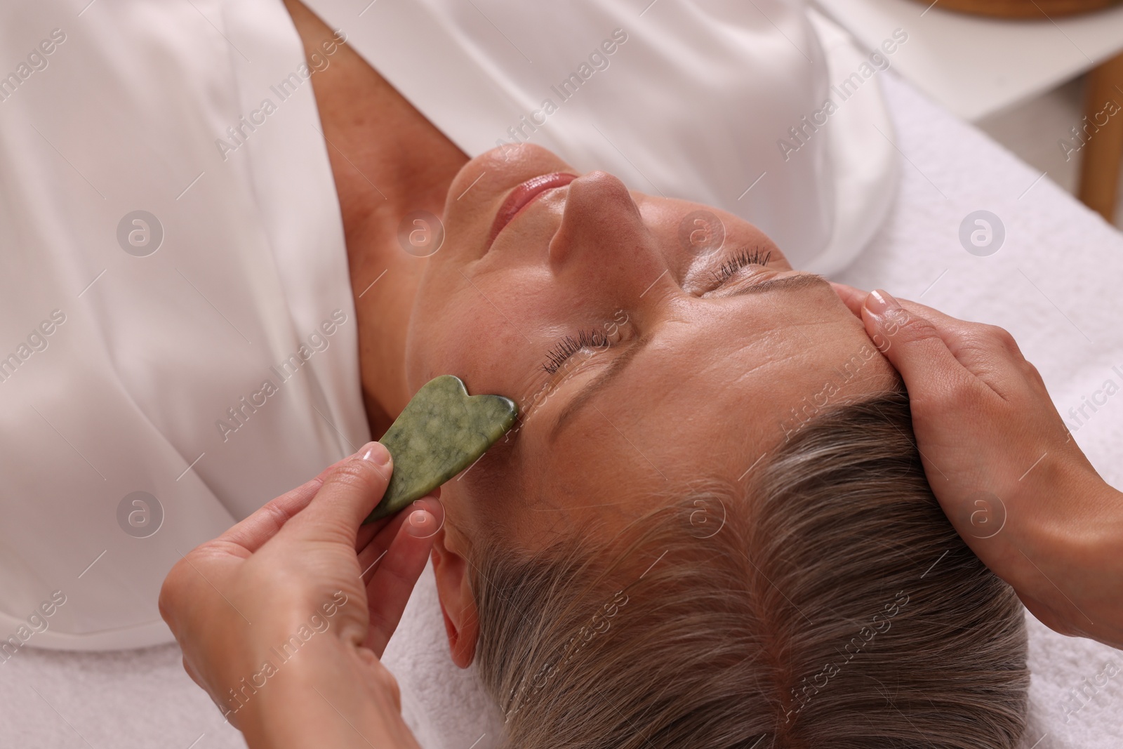 Photo of Woman receiving facial massage with jade gua sha tool in beauty salon, closeup