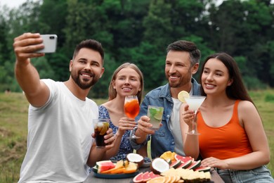 Photo of Happy friends with glasses of cocktails taking selfie at table outdoors