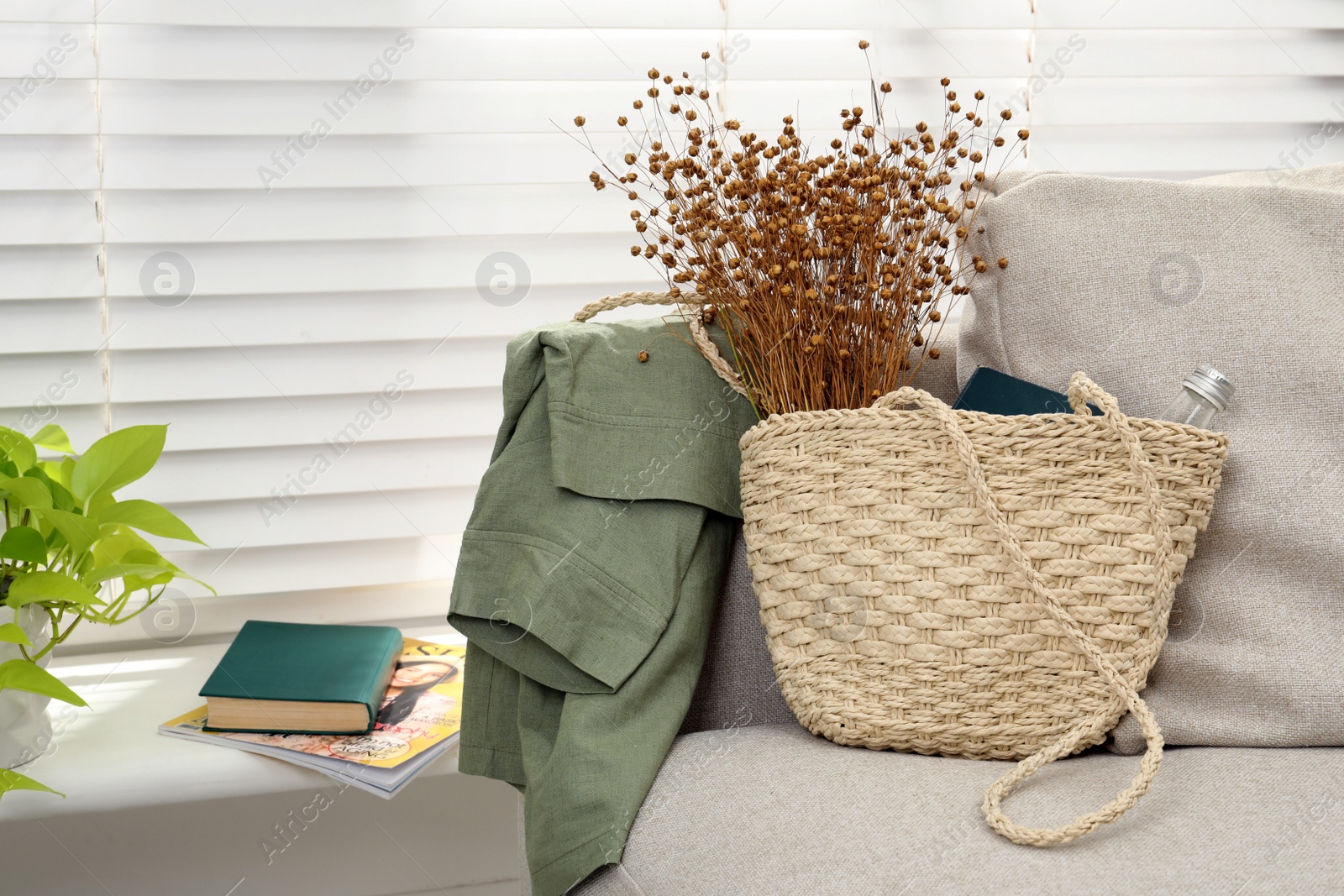 Photo of Stylish beach bag with wildflowers, bottle of water and book on sofa in room