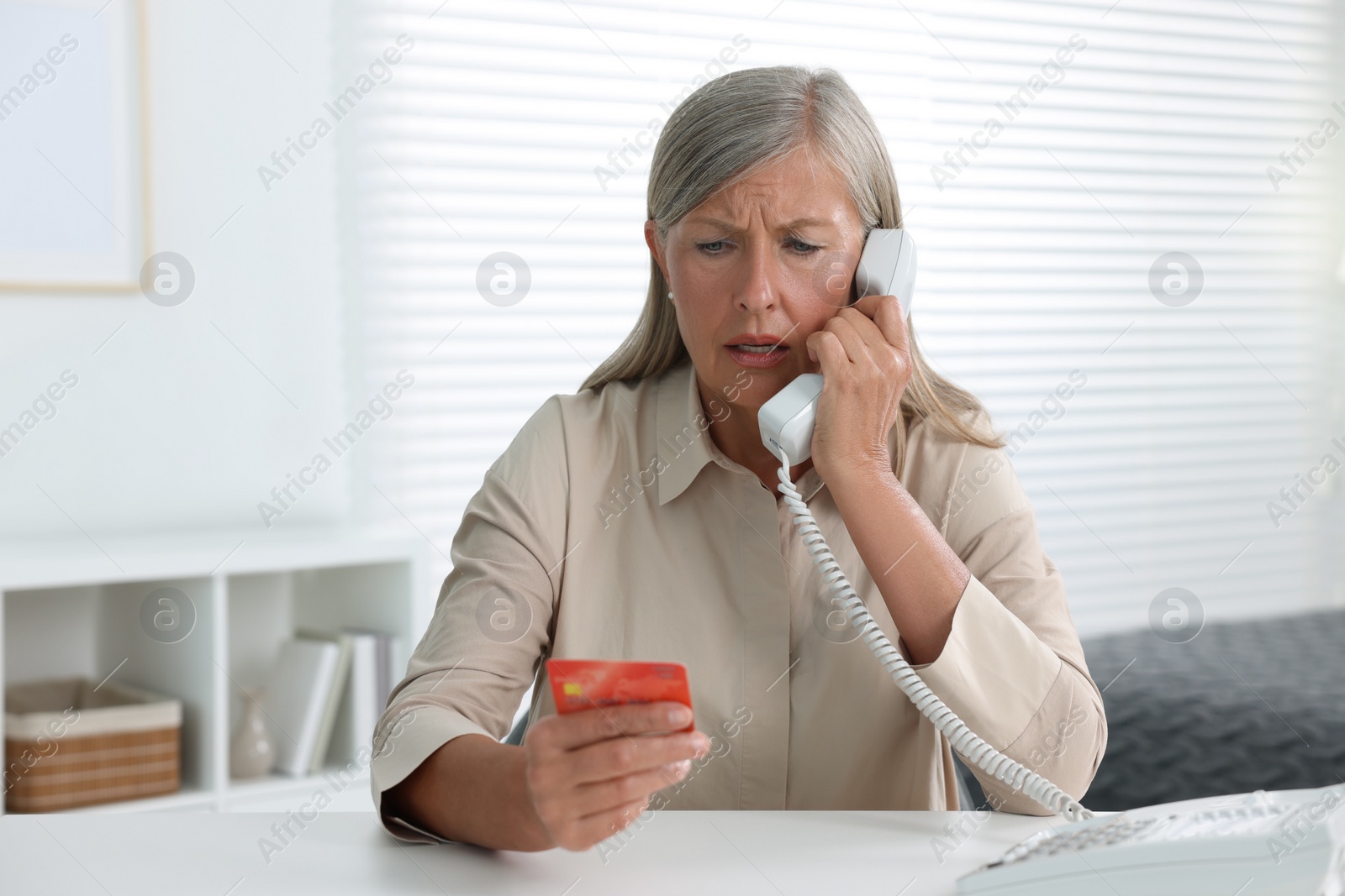 Photo of Worried woman with credit card talking on phone at table indoors. Be careful - fraud